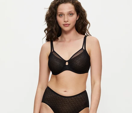 Žena s kučeravými hnedými vlasmi má na sebe čiernu podprsenku Triumph Signature Sheer Minimizer a ladiace nohavičky s vysokým pásom, pozerá do kamery.