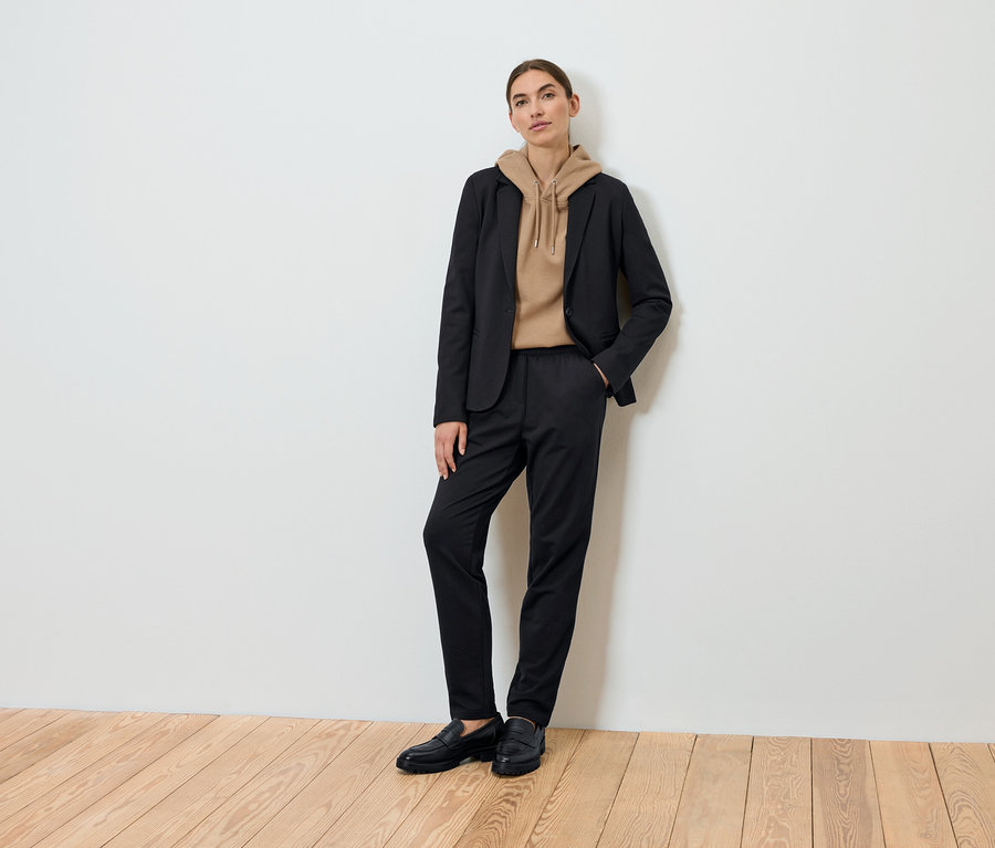 Žena pózuje v čiernom saku, hnedej mikine s kapucňou a čiernych nohaviciach.