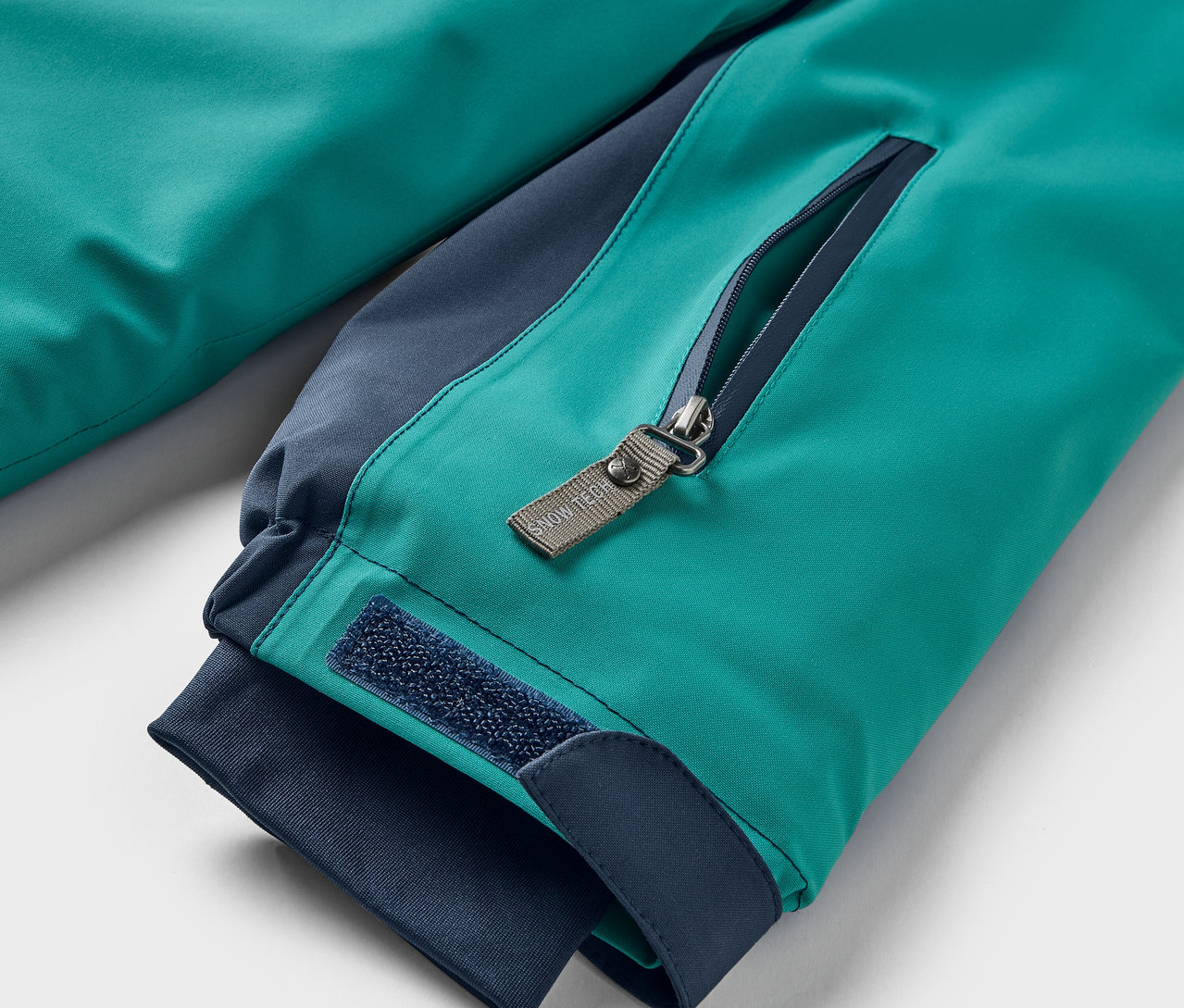 Detail tyrkysovej bundy s tmavomodrými detailmi. Z blízka je vidieť vrecko na zips a suchý zips na manžete.