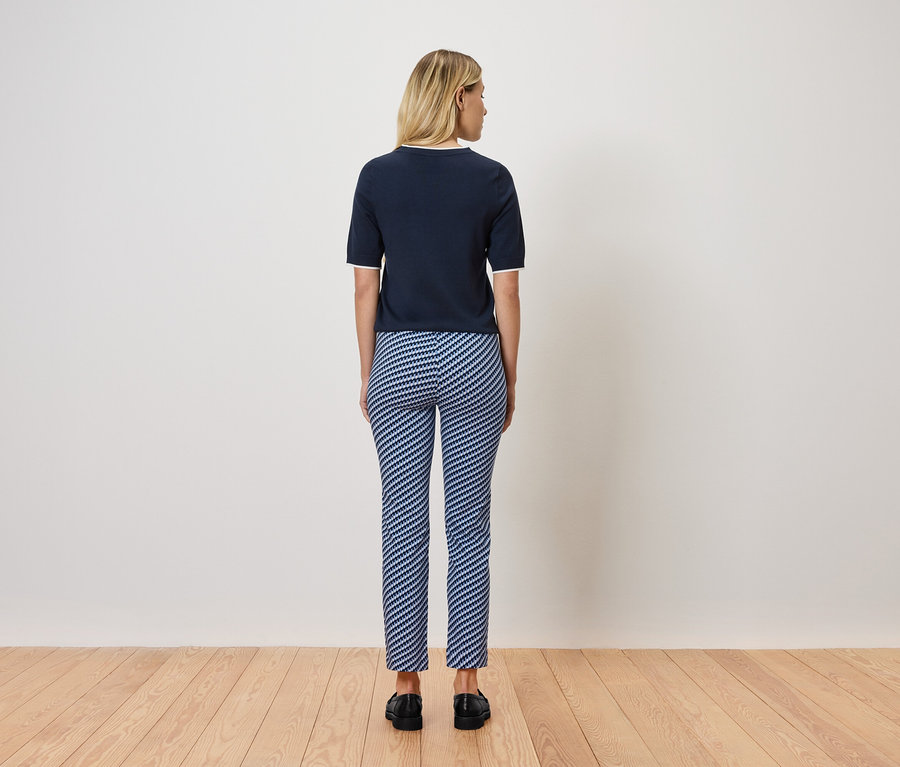 Blondínka stojí chrbtom k fotoaparátu, má na sebe tmavomodrý top s krátkym rukávom a modro-biele pruhované nohavice. Stojí na drevenej podlahe.