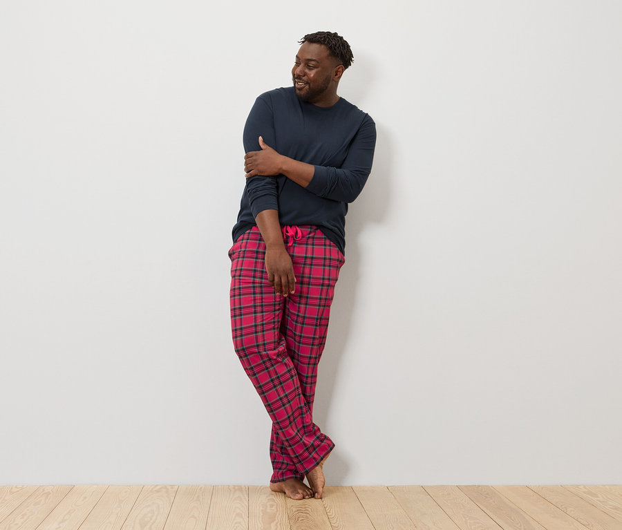 Muž opretý o stenu má na sebe tmavomodré tričko s dlhým rukávom a červené kockované pyžamové nohavice.