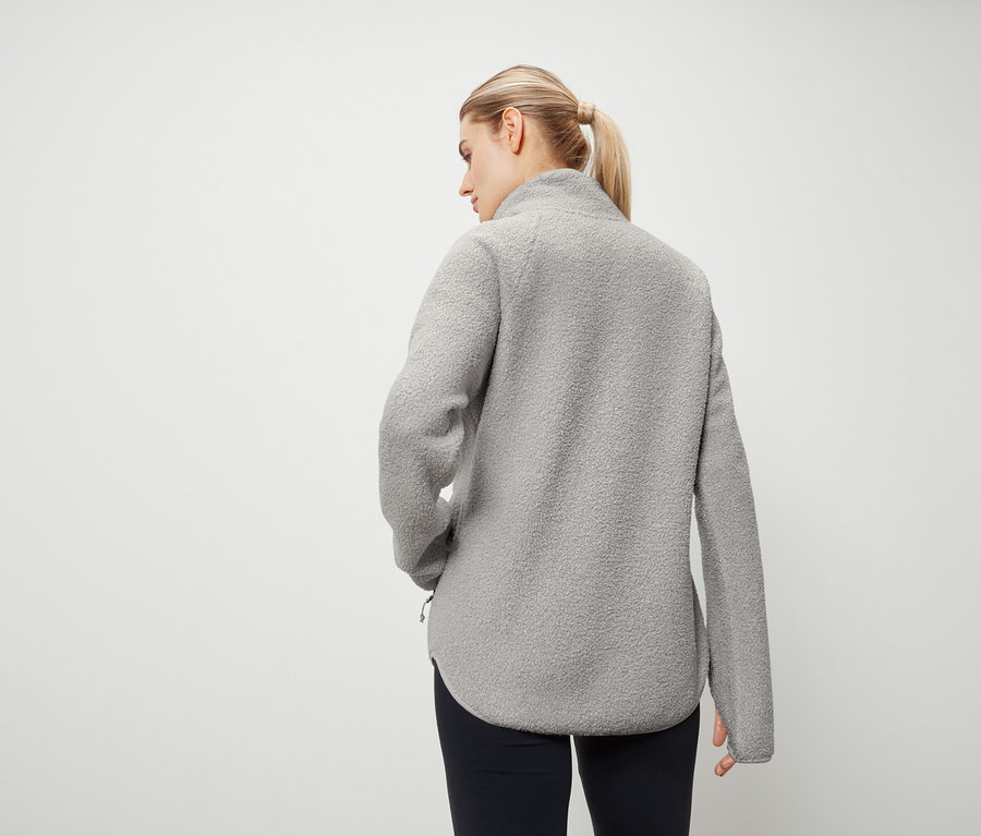 Zozadu žena s blond vlasmi stiahnutými do copu má na sebe sivú flísovú mikinu a tmavomodré nohavice.