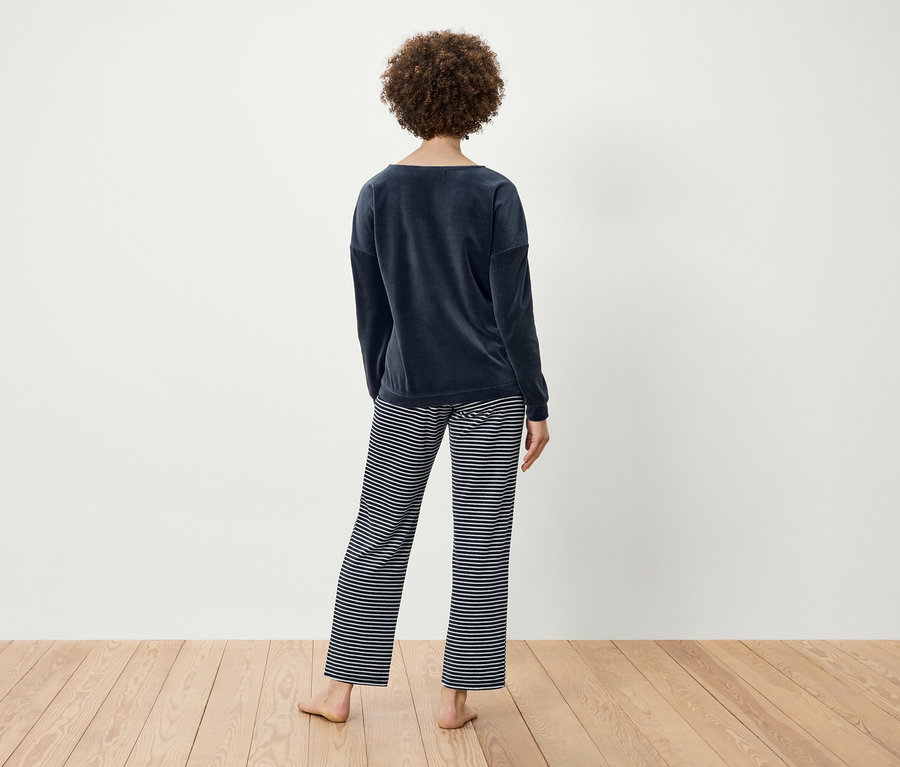 Žena s kučeravými vlasmi stojí chrbtom a má na sebe tmavomodrý zamatový top a pruhované nohavice.