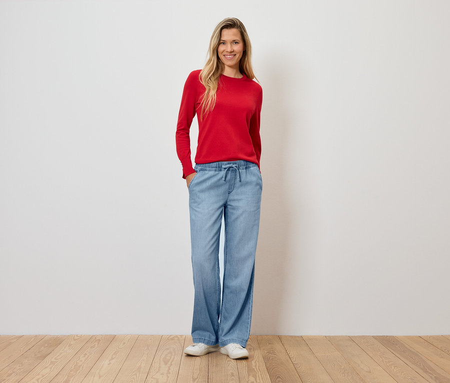 Žena v červenom svetri a širokých svetlomodrých nohaviciach stojí na drevenej podlahe.