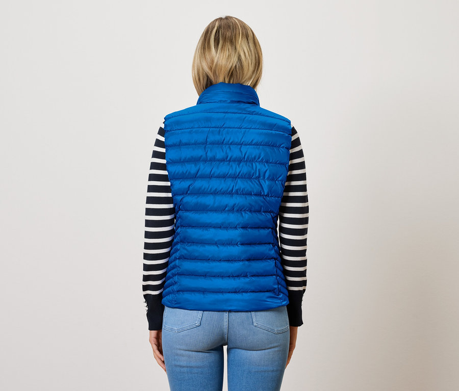 Žena chrbtom k fotoaparátu má na sebe modrú prešívanú vestu a pruhovaný sveter.