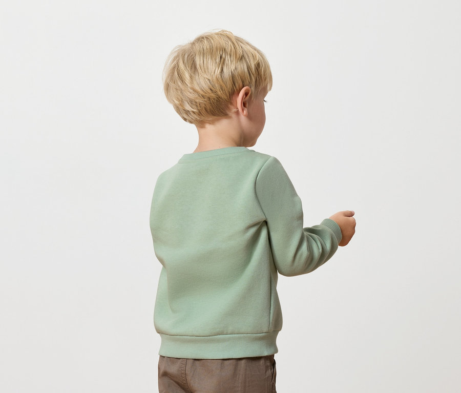 Chlapec s blond vlasmi, oblečený v zelenom svetri a hnedých nohaviciach, stojí chrbtom k fotoaparátu.