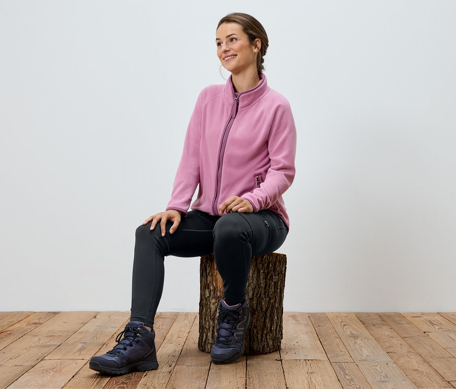 Žena sedí na drevenom kláte, má na sebe ružovú fleecovú bundu, čierne legíny a trekové topánky.