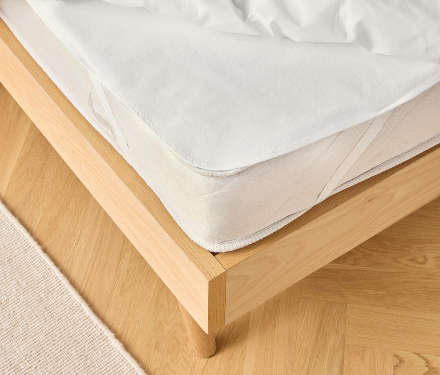 Pohľad na svetlý drevený rám postele s bielym matracom a bielou prikrývkou. Béžový koberec je čiastočne viditeľný.