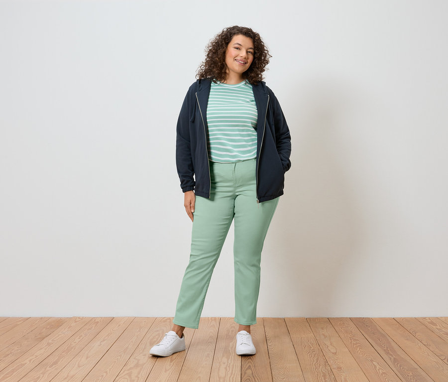 Žena s kučeravými vlasmi stojí pred stenou a má na sebe tmavomodrú bundu, pruhované tričko, zelené nohavice a biele topánky.