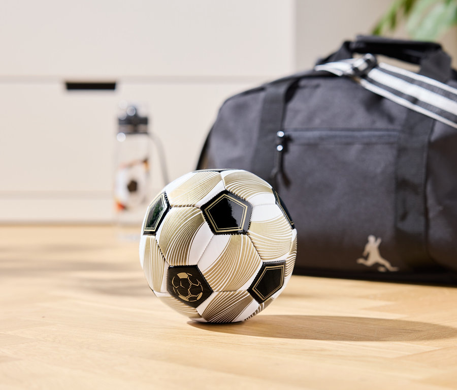 Zlatá futbalová lopta leží na drevenej podlahe, v pozadí je športová taška a fľaša na vodu.