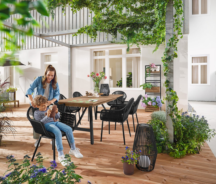 Matka a dieťa sedia pri Rozkladacom záhradnom stole s rustikálnou konštrukciou, cca 200‒250 cm. Malý a veľký Solárny lampáš s textilnou šnúrkou stojí na zemi. V pozadí je Pflanzwand mit flexibel einhängbaren Regalen »Elin«.