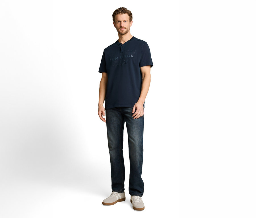 Muž v tmavomodrom tričku Henley s logom „TOM TAILOR“, tmavých džínsoch a svetlosivých teniskách.