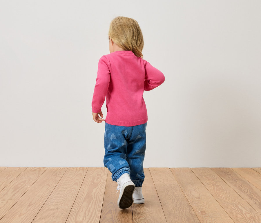 Malé blond dieťa zozadu, má na sebe ružový sveter, modré džínsy so srdiečkami a biele topánky, kráča po drevenej podlahe.