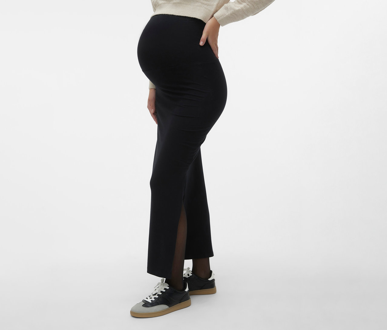 Tehotná žena nosí maxi sukňu MAMALICIOUS »MLEMMA TUBE« s bočným rozparkom, tmavé pančuchy a tenisky na neutrálnom pozadí.