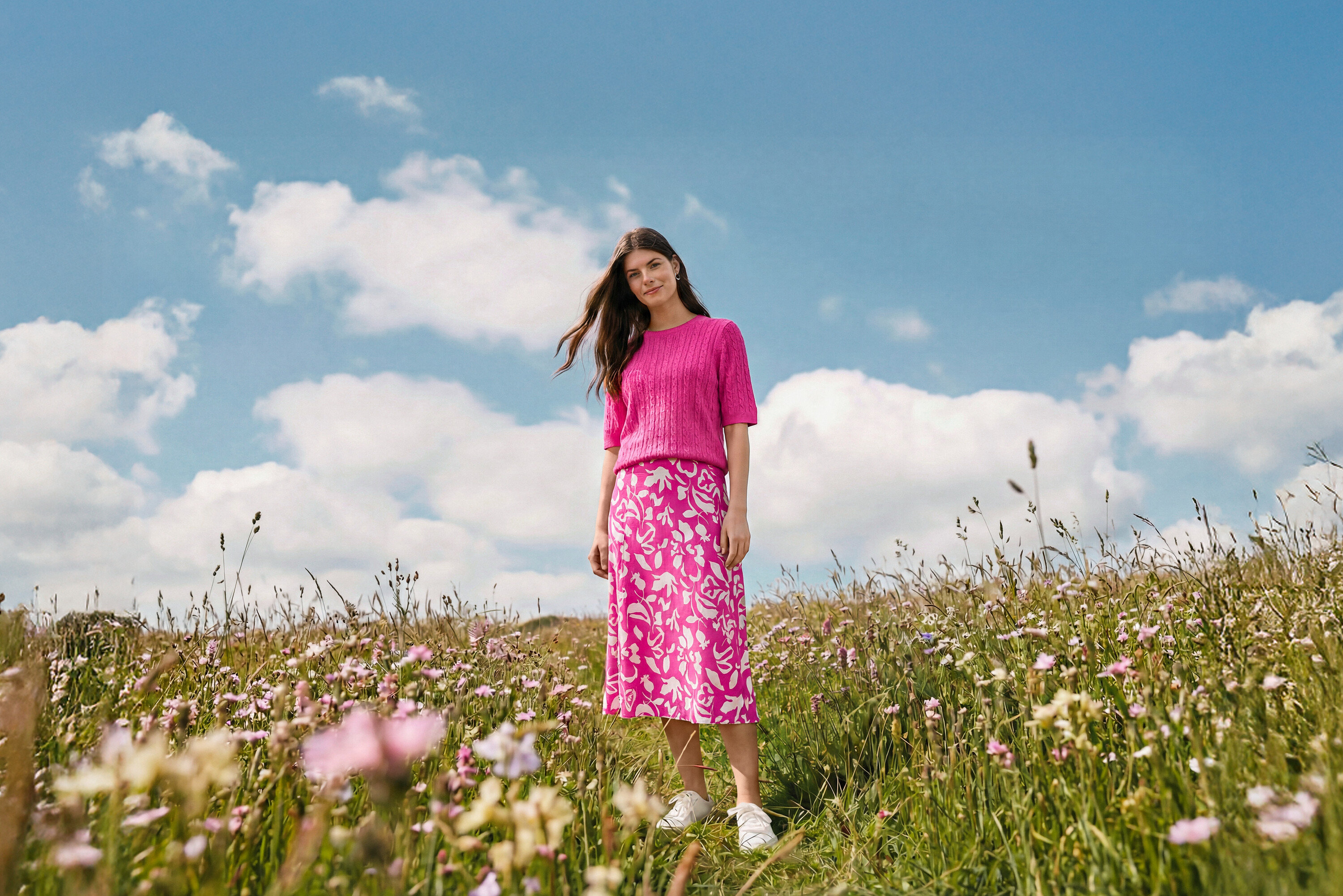 Žena stojí na lúke a má na sebe bedruckter midi-rock, kruhové náušnice Wave a kožené tenisky.