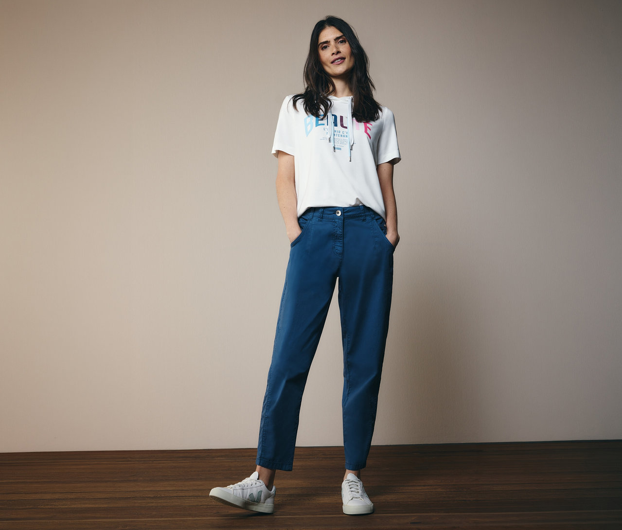 Žena s tričkom s kapucňou CECIL a modrými nohavicami CECIL Barrel »Scarlett« stojí s rukami vo vreckách pred kamerou.