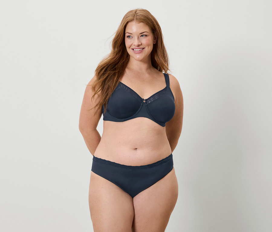 Žena s dlhými ryšavými vlasmi má na sebe tmavomodrú podprsenku a nohavičky a usmieva sa.