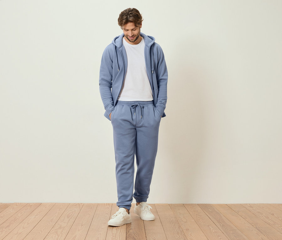 Muž stojí s rukami vo vreckách v svetlomodrej teplákovej súprave a bielych teniskách.