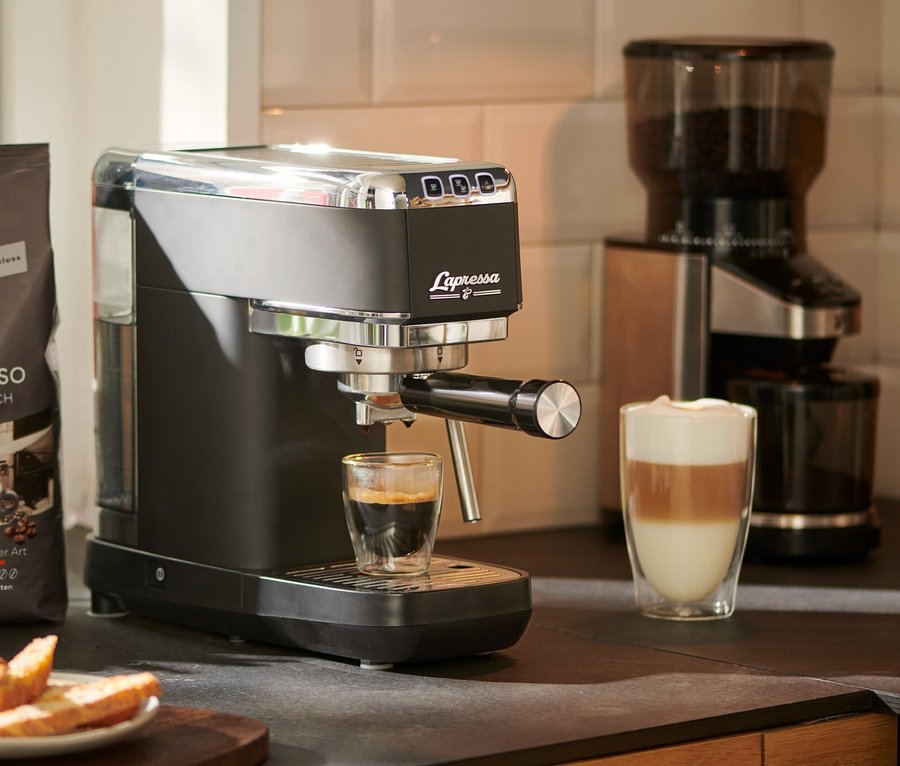 Espresso tečie z pákového kávovaru »Lapressa«, čierny, do sklenice. V pozadí je mlynček na kávu a balíček kávy.