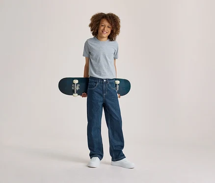 Usmiaty chlapec s kučeravými vlasmi v sivom tričku, modrých džínsoch a bielych teniskách, držiaci za chrbtom tmavomodrý skateboard.
