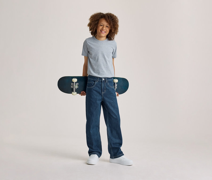 Usmiaty chlapec s kučeravými vlasmi v sivom tričku, modrých džínsoch a bielych teniskách, držiaci za chrbtom tmavomodrý skateboard.