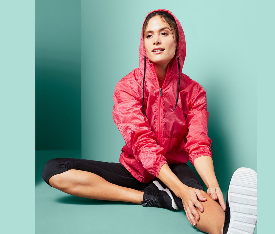 Žena sedí na zemi a preťahuje sa v ľahkej športovej bundě.