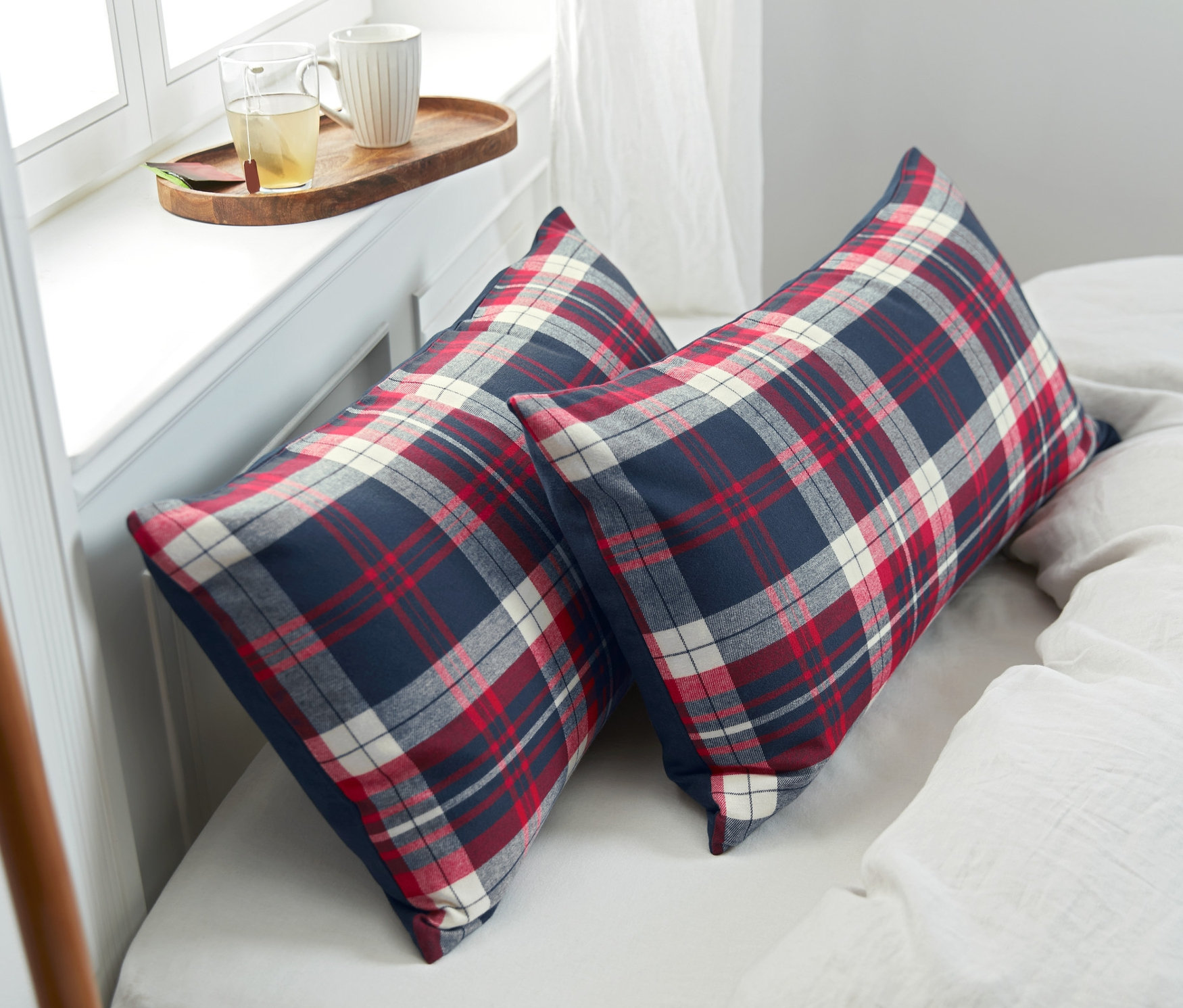 Dve flanelové obliečky na vankúše s červeným, bielym a modrým kockovaným vzorom ležia na posteli. Vedľa nich je sivá deka a podnos so šálkami.