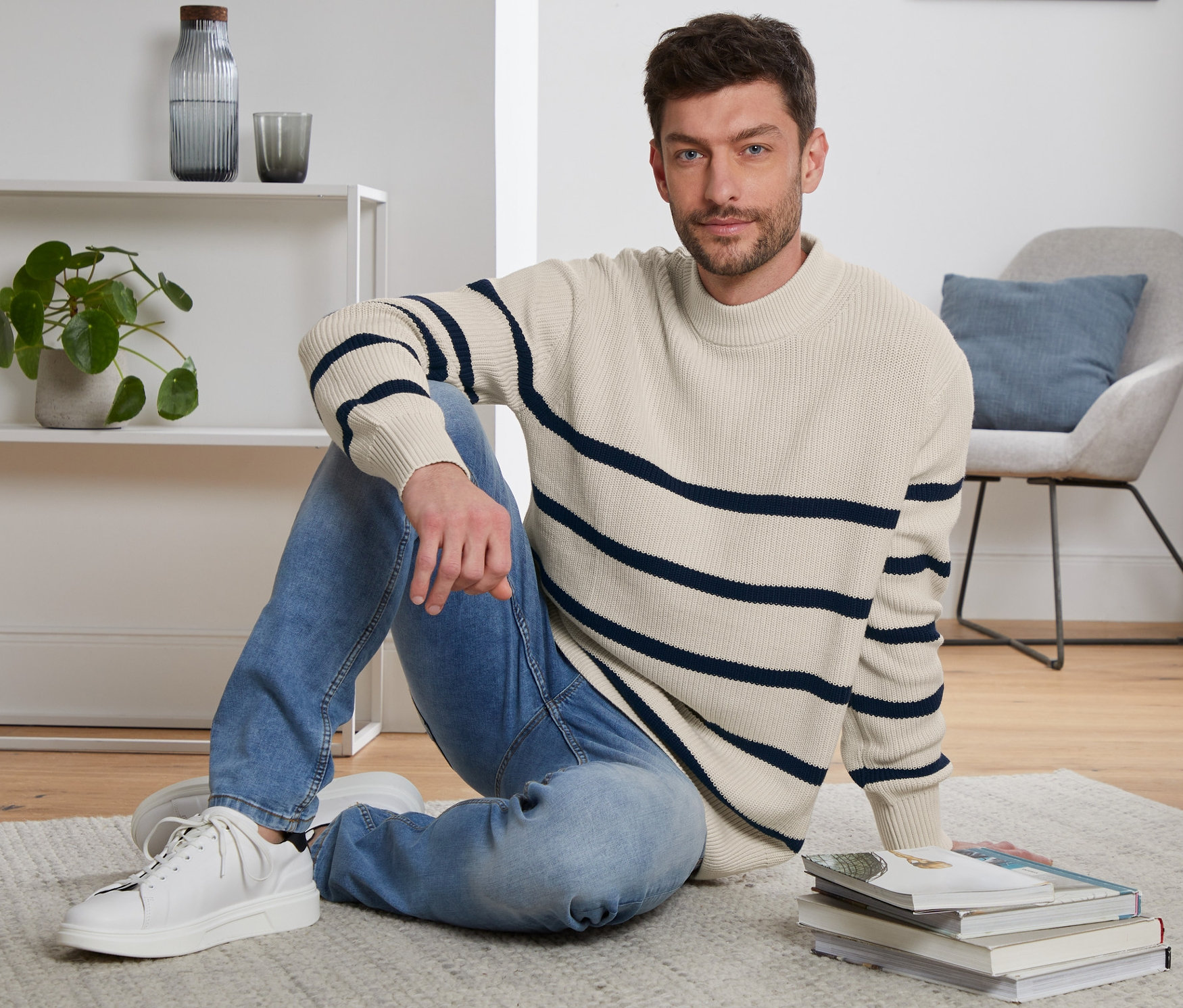 Muž sedí na podlahe na koberci a má na sebe pletený pulóver.