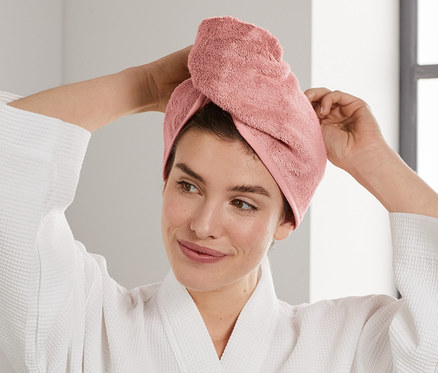 Žena si upravuje ružový uterákový turban na hlave. Oblečená v bielom župane stojí v kúpeľni.
