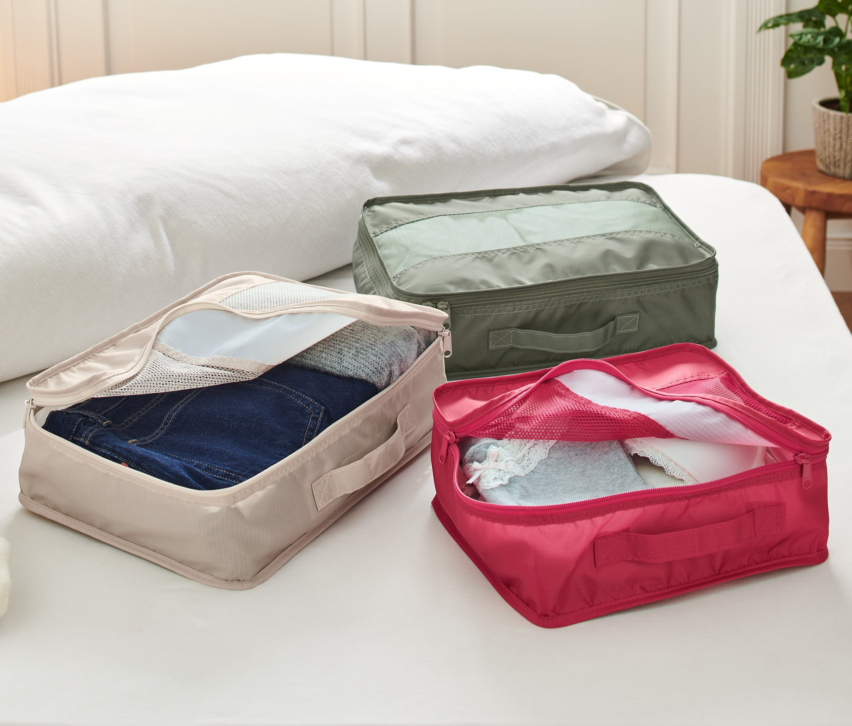 Na posteli ležia tri cestovné organizéry, každý s inou farbou a obsahom.