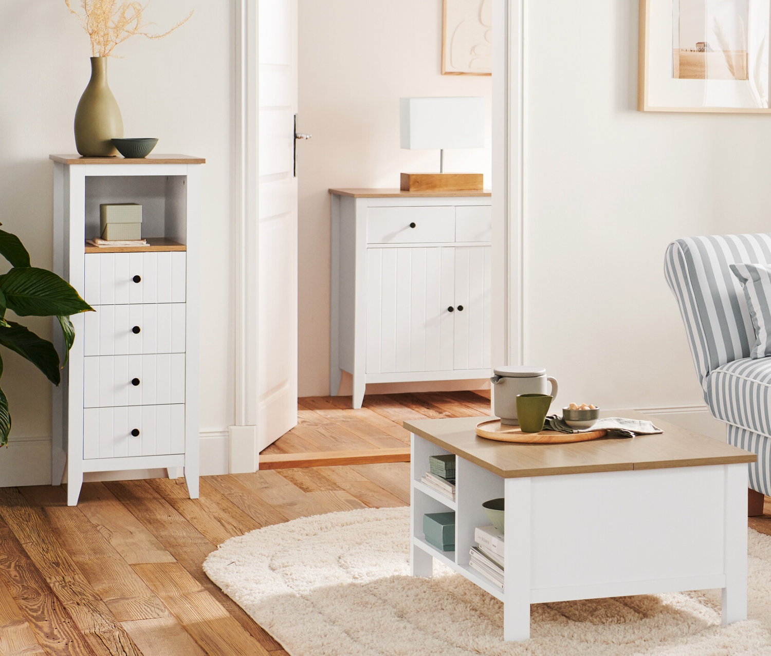 V obývacej izbe stojí drevená stojacia lampa »Trojnožka« vedľa pohovky a konferenčného stolíka s funkciou sklápania. V pozadí je komoda so zásuvkami a stolová lampa s dreveným podstavcom.