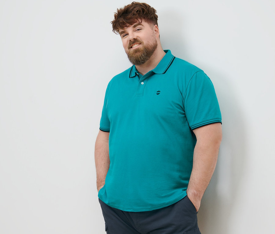 Muž s hnedými vlasmi stojí a má ruky vo vreckách cargo šortiek námorníckej modrej farby. Má na sebe akvamarínovú piké polokošeľu.