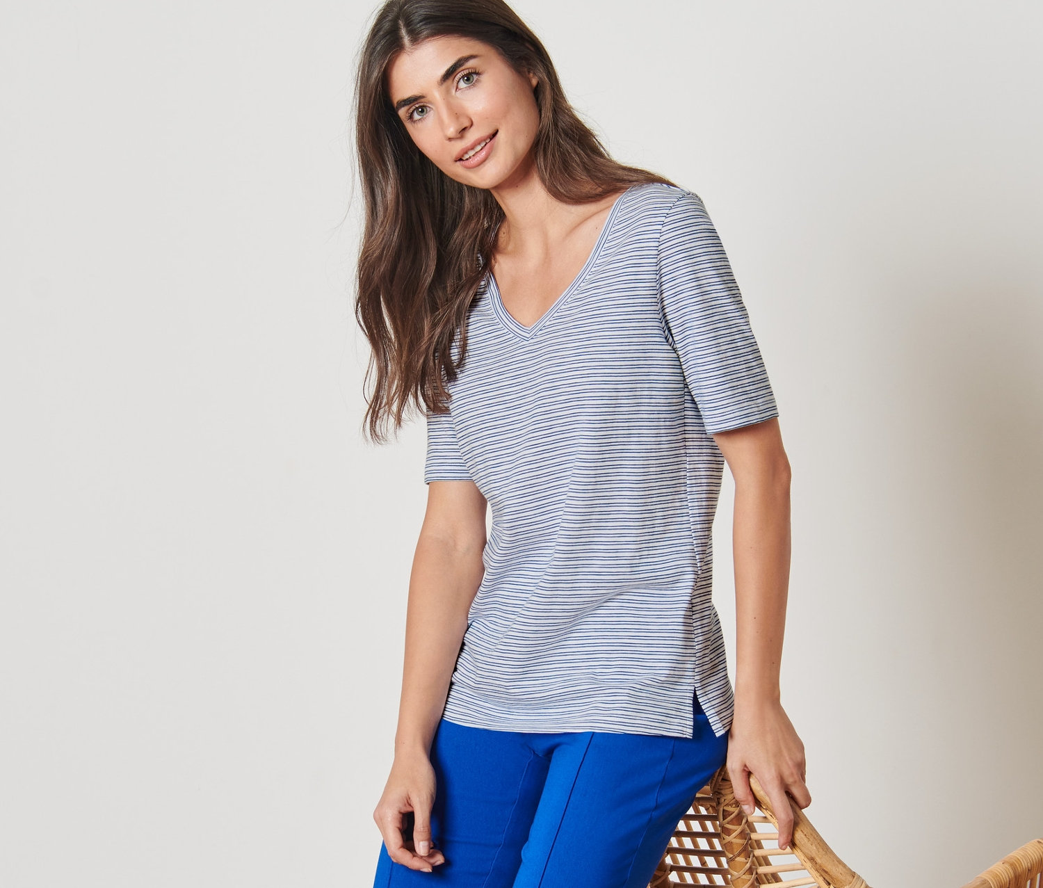 Žena opierajúca sa o prútené kreslo má na sebe prúžkované tričko a modré nohavice.