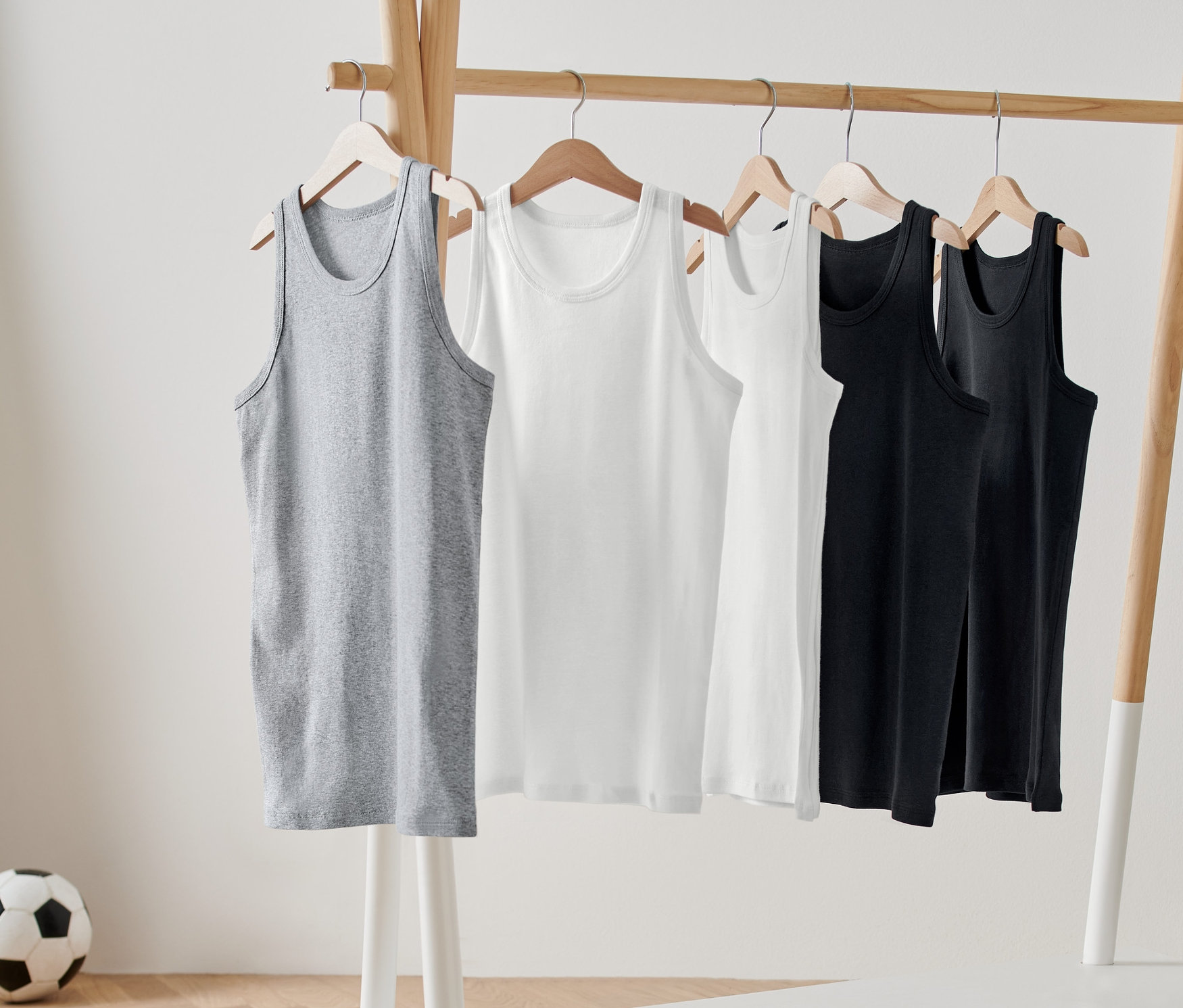 Päť detských tielok v sivej, bielej a čiernej farbe visí na drevenom vešiaku. V pozadí je vidieť futbalovú loptu.