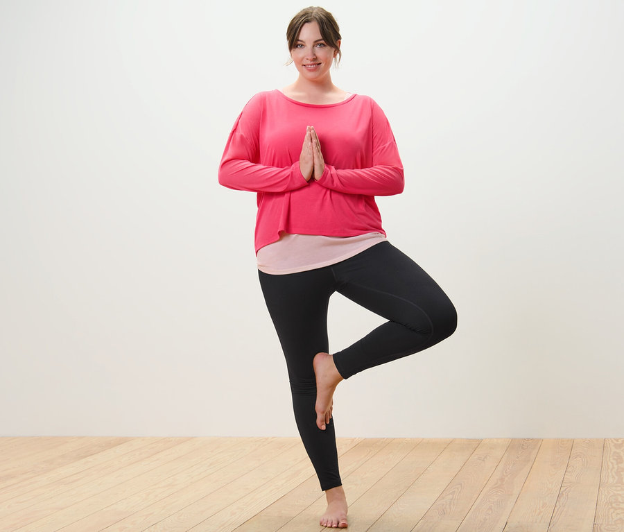 Žena v ružovom športovom tričku 2 v 1 a čiernych športových legínach stojí v pozícii stromu.