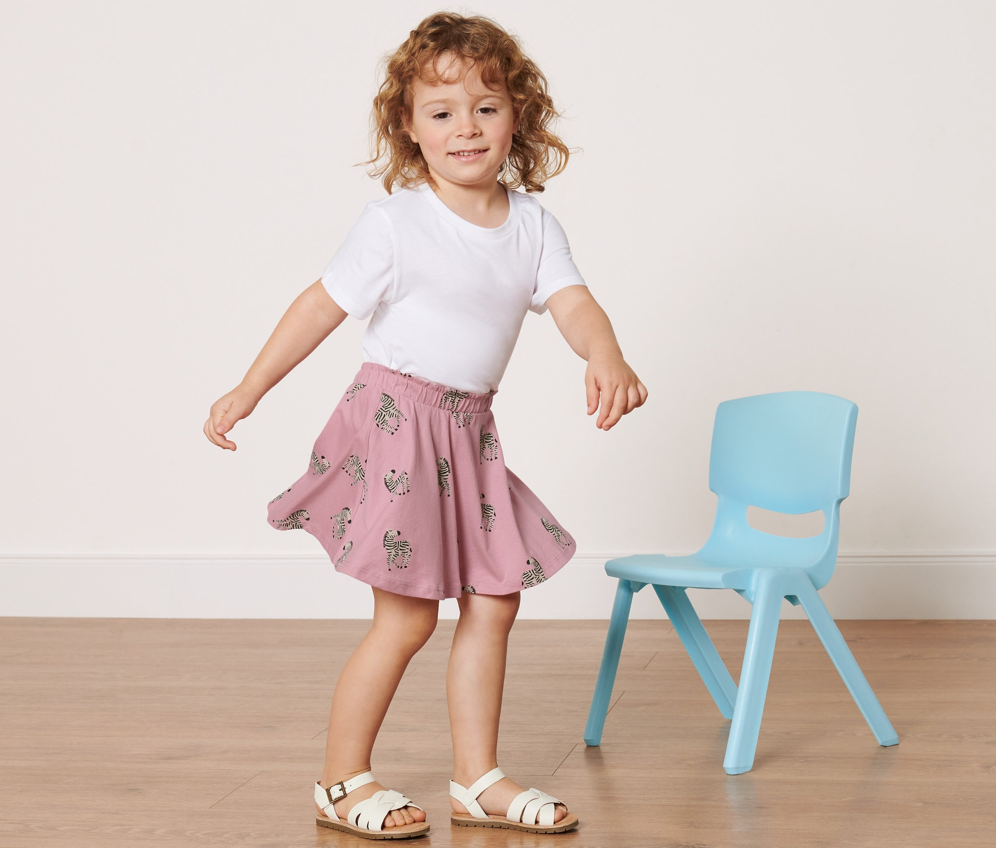 Dievča v bielom tričku a Detská džersejová sukňa s nohavicami stojí vedľa modrej stoličky.