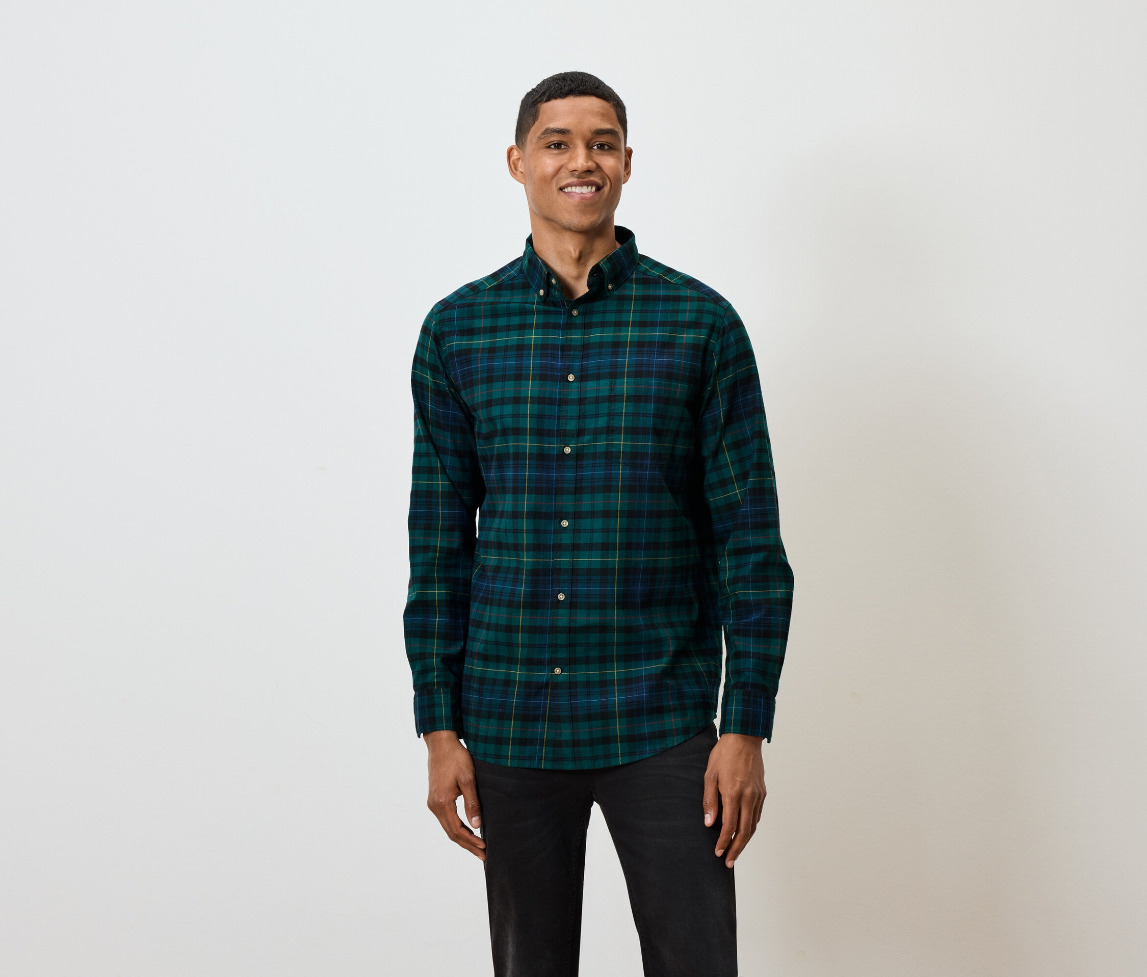 Muž s krátkymi vlasmi pózuje na bielom pozadí a má na sebe zeleno-modrú kockovanú košeľu s dlhým rukávom a čierne džínsy.