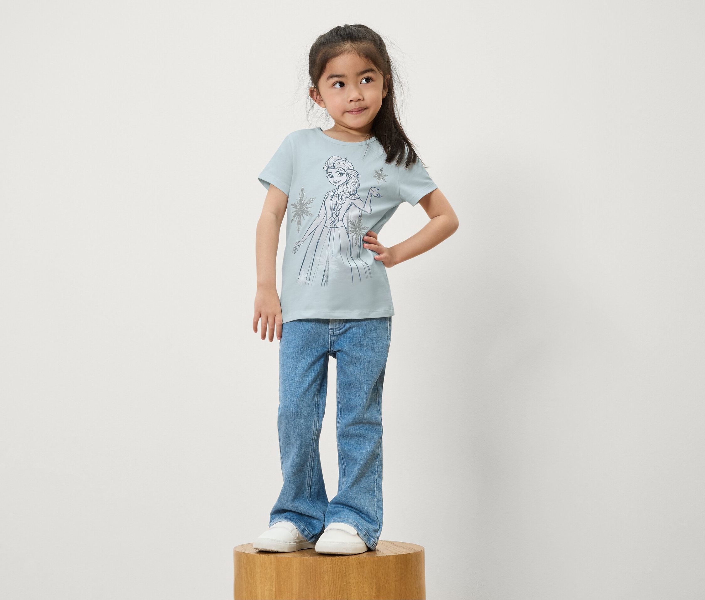 Dievča stojí na drevenom podstavci. Má na sebe modré džínsy a svetlomodré tričko s potlačou Elsy z Ľadového kráľovstva.