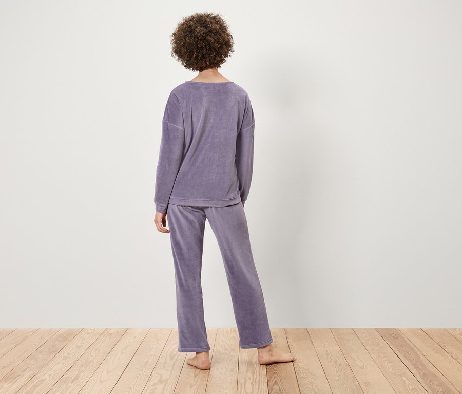 Žena s kučeravými vlasmi stojí chrbtom, má na sebe fialové zamatové pyžamo na svetlom drevenom povrchu.