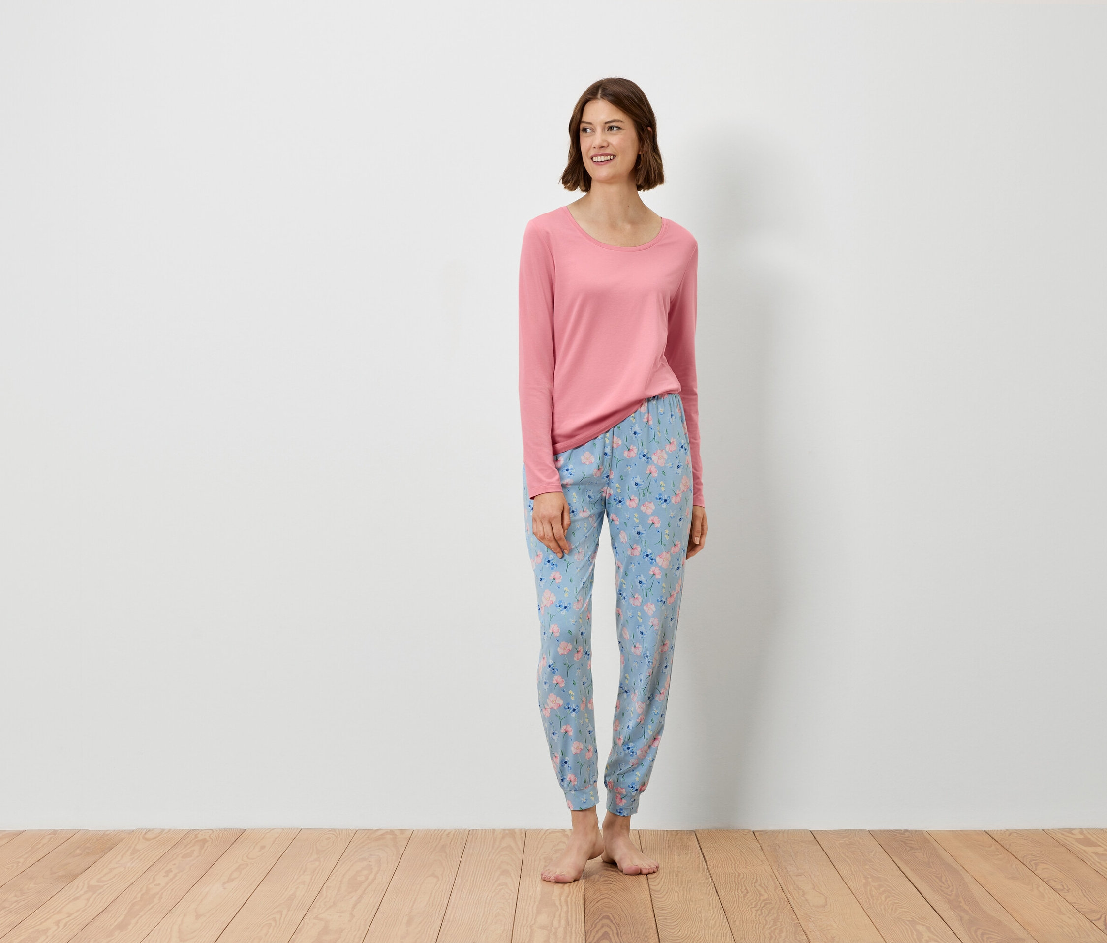 Žena stojí a má na sebe ružový top s dlhým rukávom a modré nohavice s kvetinovým vzorom.