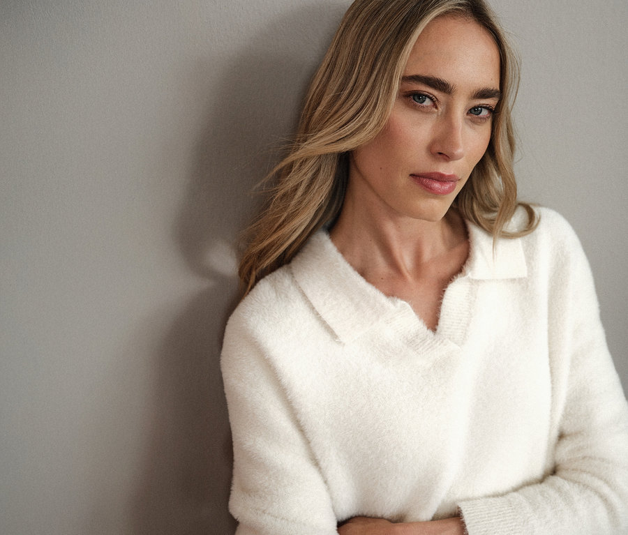 Žena s blond vlasmi v bielom pletenom pulóvri s polo golierom Street One sa opiera o sivú stenu.