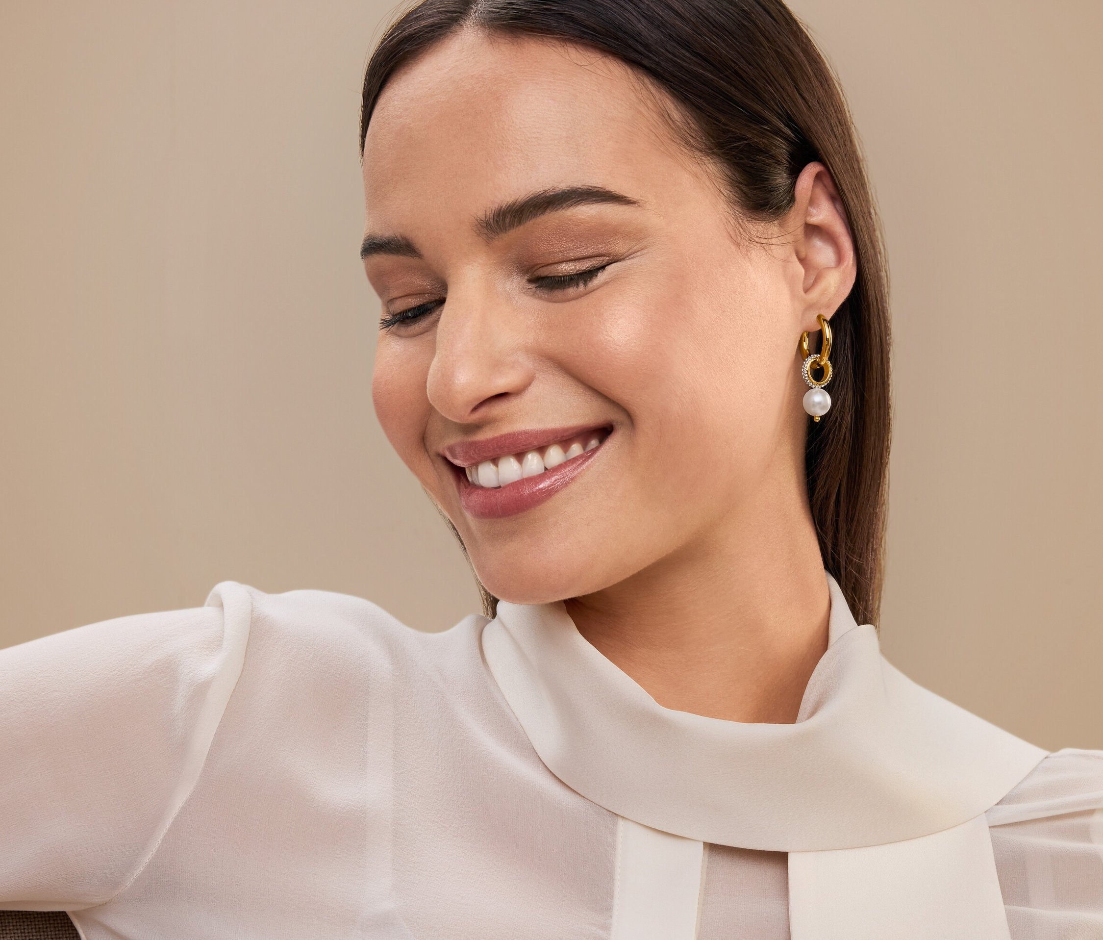 Usmievajúca sa žena s hnedými vlasmi a zlatými náušnicami s perlou, má na sebe bielu blúzku.
