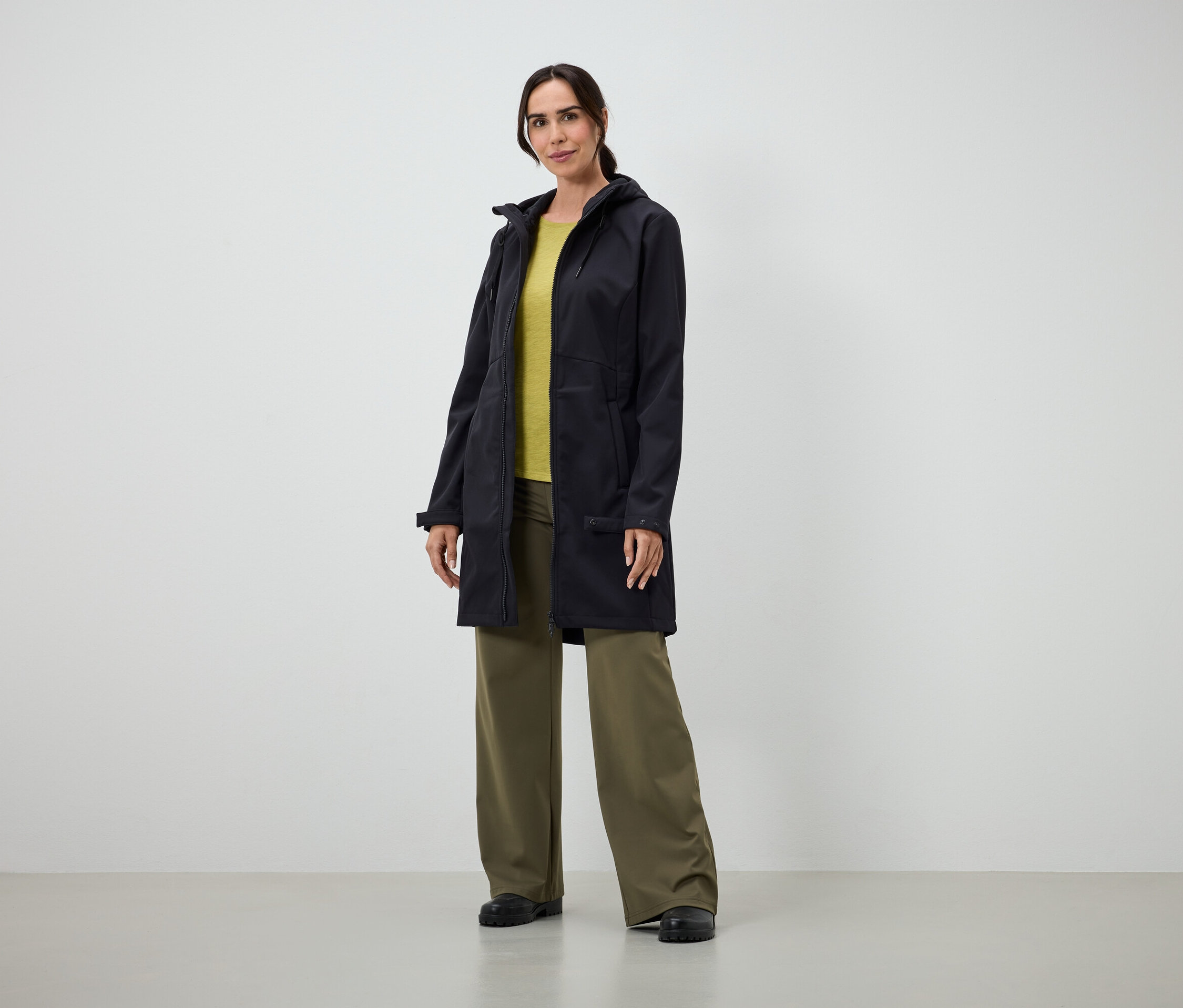 Žena v dlhom čiernom kabáte, žltom tričku a širokých zelených nohaviciach stojí v interiéri.