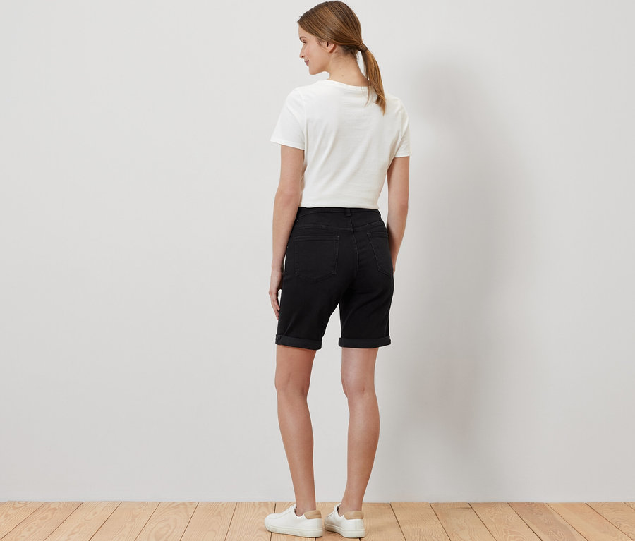 Žena stojí chrbtom a má na sebe džínsové bermudy – strih »Lea«, čierny denim, biele tričko a tenisky.