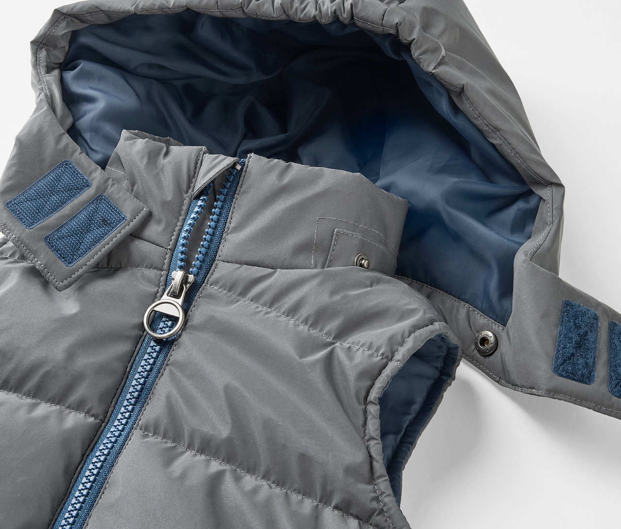 Detail šedej prešívanej vesty s kapucňou. Reflexná detská prešívaná vesta má modrý zips a modrú vnútornú stranu kapucne.