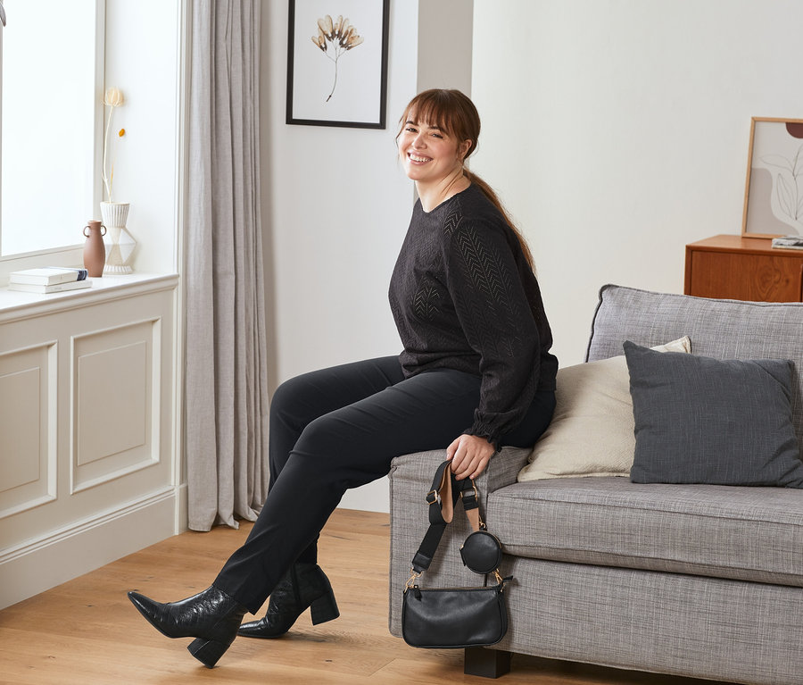 Žena sedí na pohovke. Má na sebe kožené topánky, elastické nohavice tmavočervené a drží malú taštičku červenú.