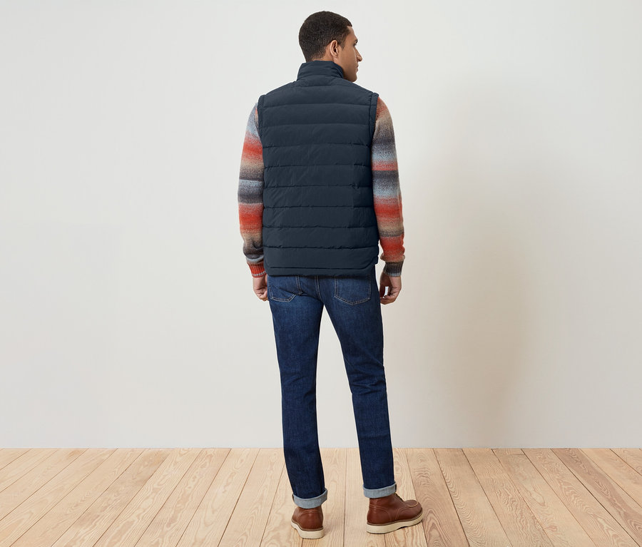 Muž chrbtom ku kamere má na sebe tmavomodrú vestu, farebný sveter, džínsy a hnedé topánky.