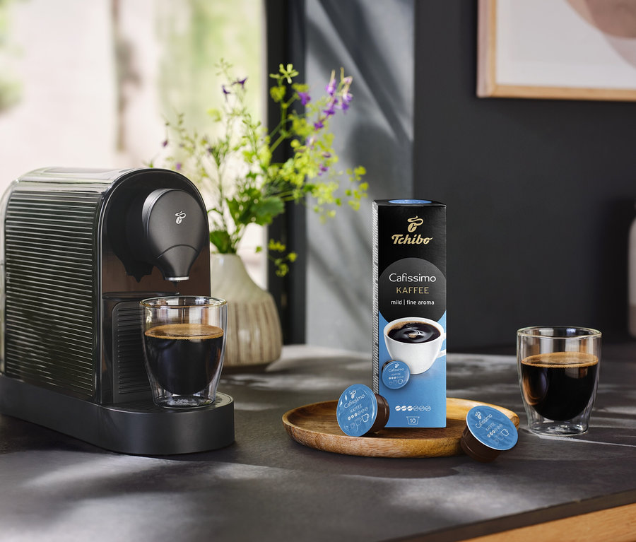 Kávovar stojí vedľa pohára kávy a balenia 10 kapsúl Káva jemná.