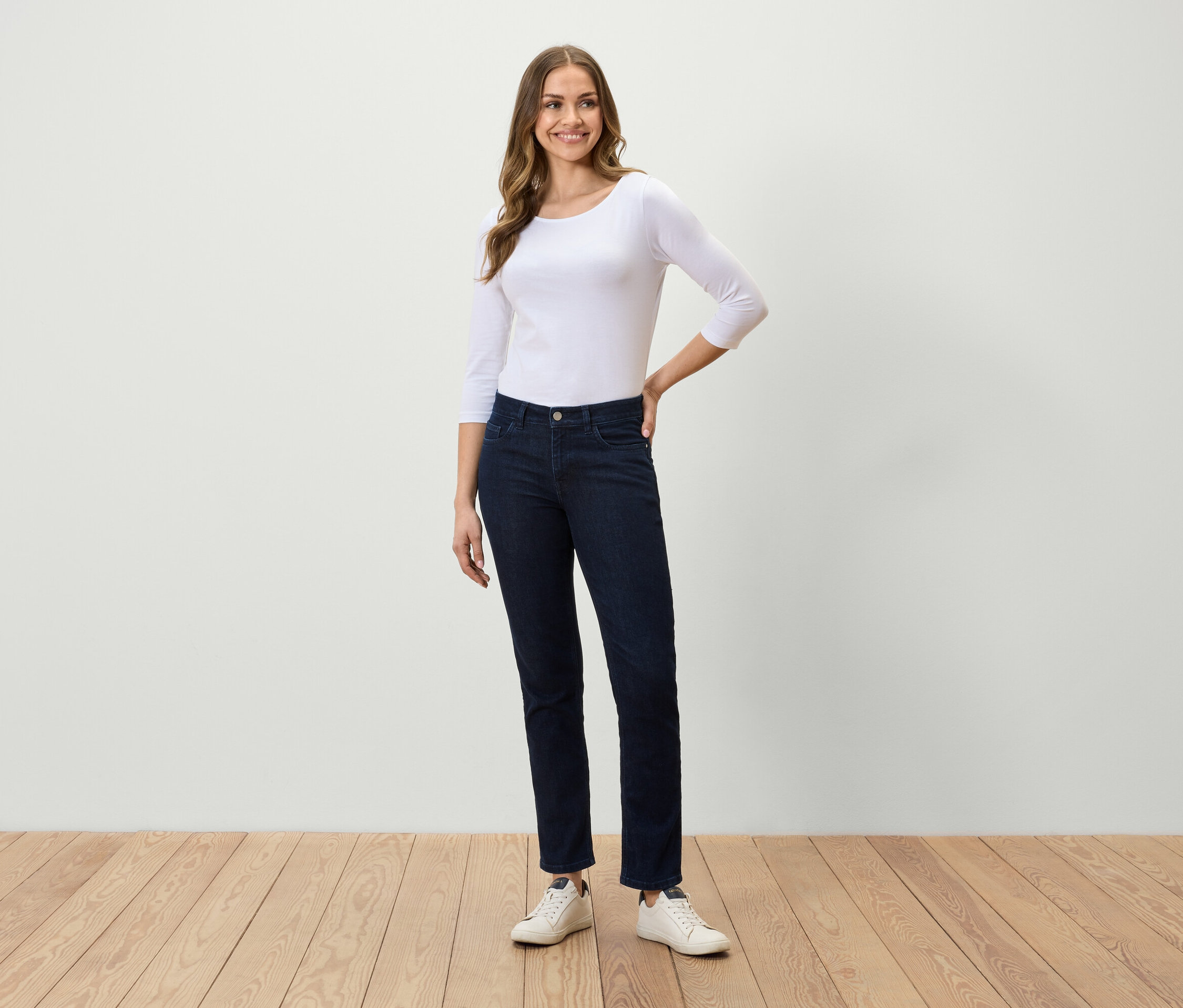 Žena stojaca na drevenej podlahe má na sebe biele tričko s trojštvrťovým rukávom, tmavomodré džínsy a biele tenisky.