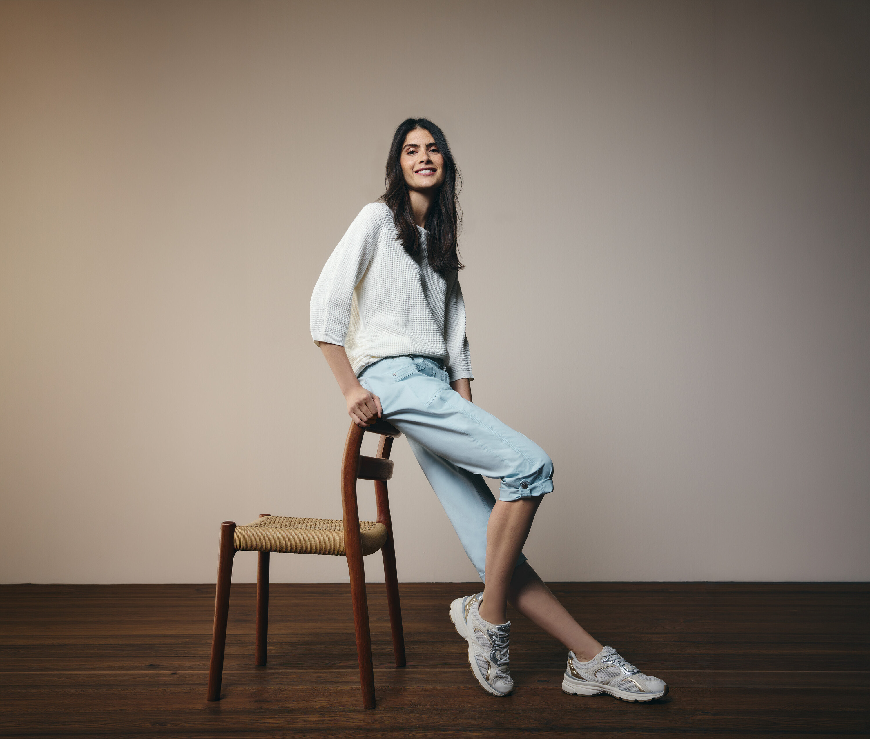 Žena sa opiera o stoličku, má na sebe svetlomodré 3/4 nohavice Cecil »New York« a biely sveter.